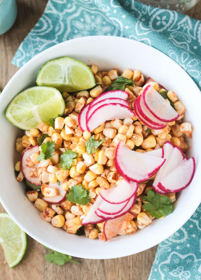 Raw Corn Radish Salad in a white bowl garnished with extra thinly sliced radishes, lime wedges, and cilantro leaves.