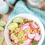Corn Radish Salad in a white bowl garnished with cilantro leaves and lime wedges. Extra lime slices on the side with blue salt and pepper shakers in the background.