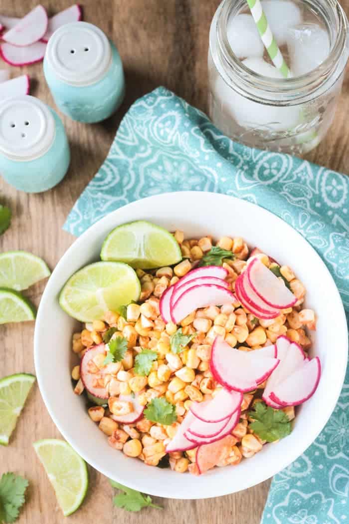 Corn Radish Salad in a white bowl garnished with cilantro leaves and lime wedges. Extra lime slices on the side with blue salt and pepper shakers in the background.
