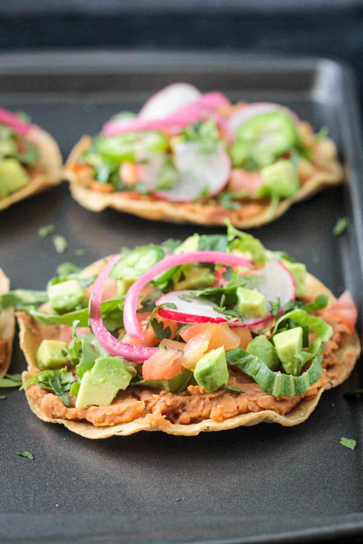 Close up of a crispy tostada shell topped with refried beans, avocado, and pickled onion.
