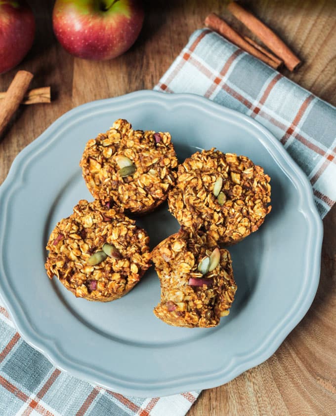 Plate of 4 Apple Pumpkin Pie Baked Oatmeal muffins. One has been bitten into. Two red apples behind the plate next to some cinnamon sticks.