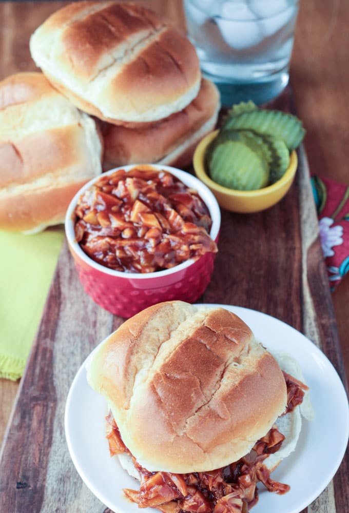 BBQ Jackfruit sandwich in front a bowl of bbq jackfruit. Small yellow bowl of pickles and a stack of burger buns behind next to a glass of ice water.