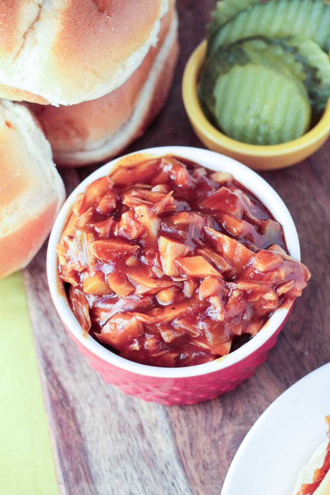 Red bowl of bbq jackfruit next to a small yellow bowl full of pickle slices. Burger buns stacked nearby.