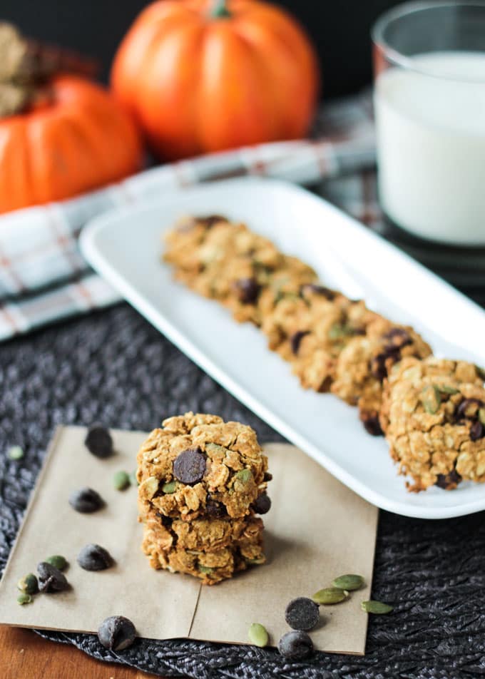 Small stack Pumpkin Oatmeal Chocolate Chip Cookies in front of more cookies lined up on a white rectangular platter. Glass of milk in the background.