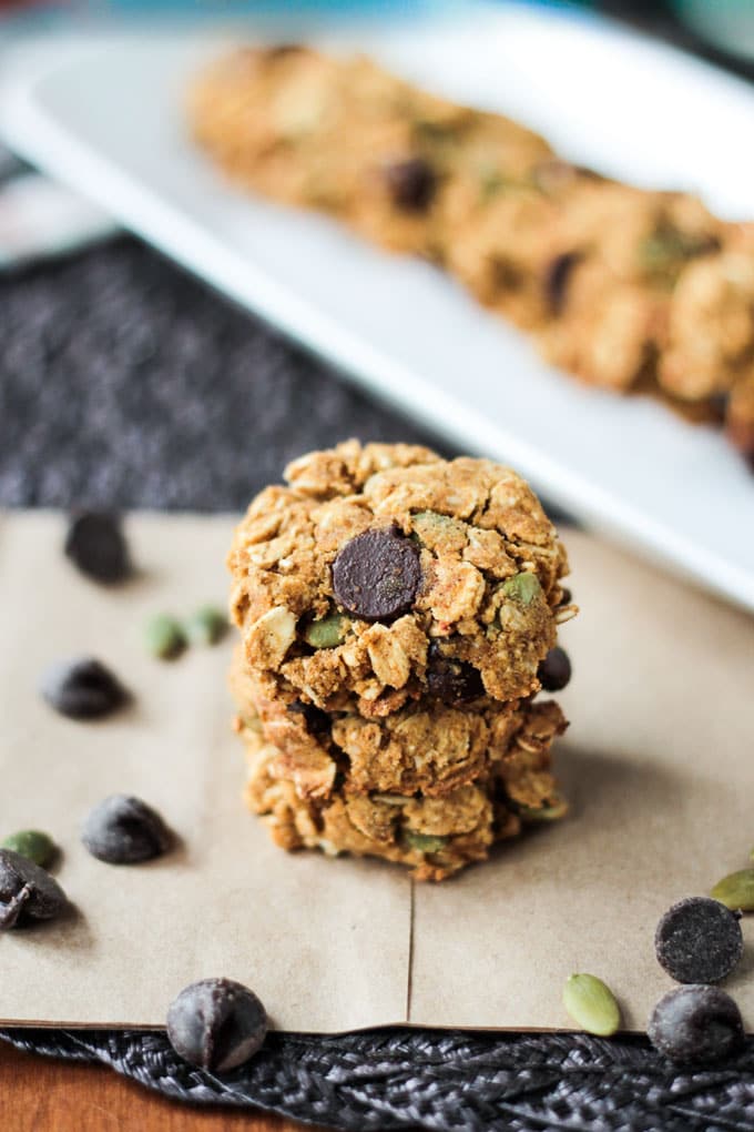 Crunchy Gluten Free Pumpkin Oatmeal Chocolate Chip Cookies - these healthy, oil free, vegan cookies are addictive! Super crunchy & filled with nutritious ingredients and fall flavors. Eat them for dessert, snack, or even breakfast! I dare you to eat just one! Stack of three Pumpkin Oatmeal Chocolate Chip Cookies. A white dish with a row of cookies blurred out in the background.