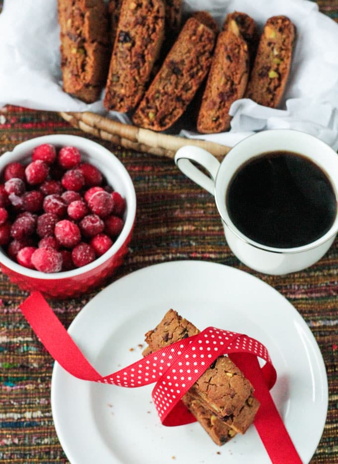 Basket Cranberry Pistachio Biscotti next to a bowl of fresh cranberries, cup of coffee, and a plate of three stacked biscotti tied in a red ribbon.