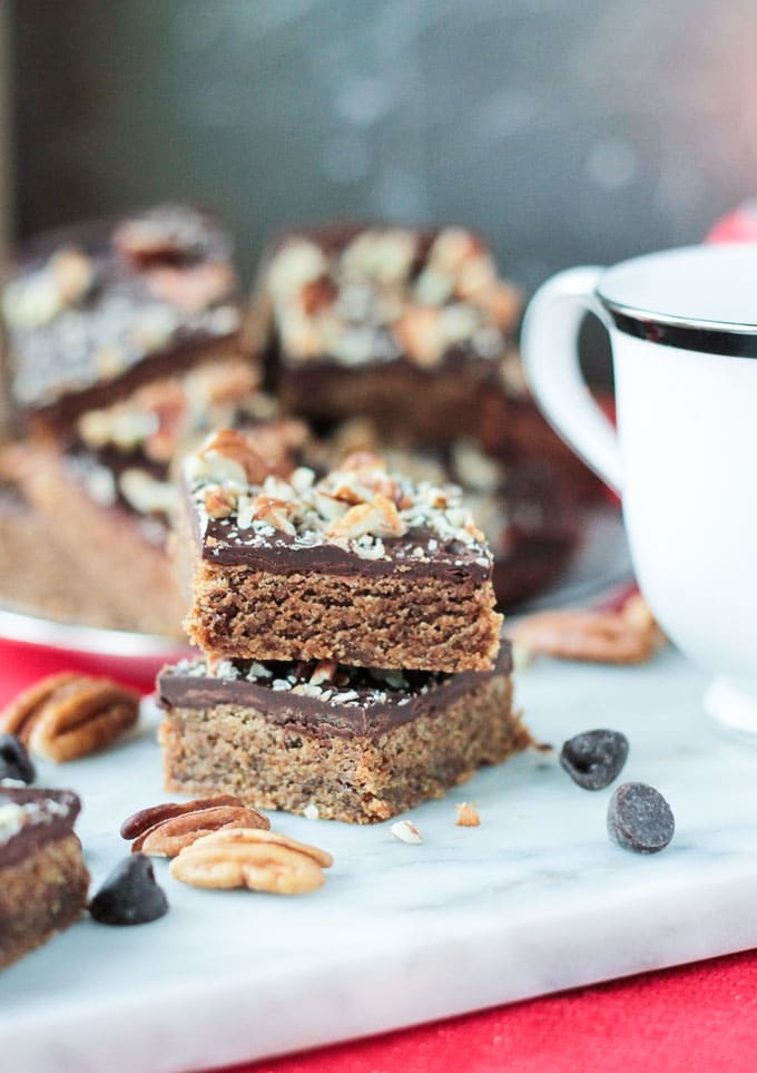 2 Vegan Toffee Bars stacked on a white platter with pecans and chocolate chips scattered around them.