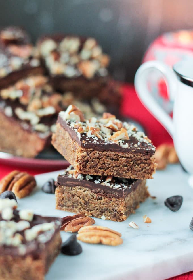 Two Vegan Toffee Bars stacked in front a plate full of more bars. Pecans and chocolate chips scattered about.