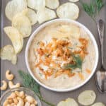 Bowl of vegan french onion dip on a tray surrounded by potato chips and a small bowl of cashews.