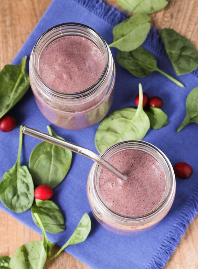 Overhead view of two Coconut Water Smoothies in glasses on a blue dish cloth surrounded by fresh spinach leaves and cranberries.