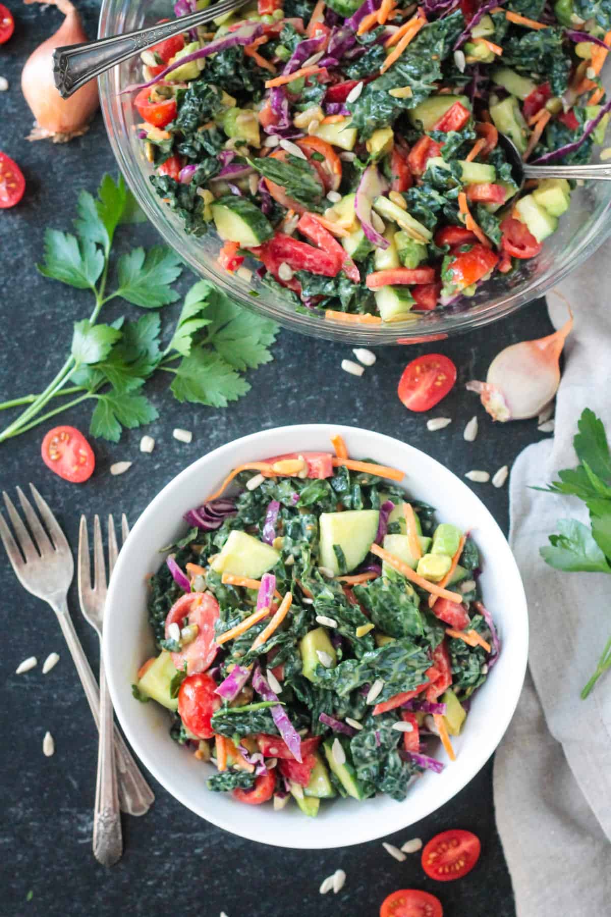 A serving of kale salad in a white bowl on a table with a large bowl of more salad and two forks.