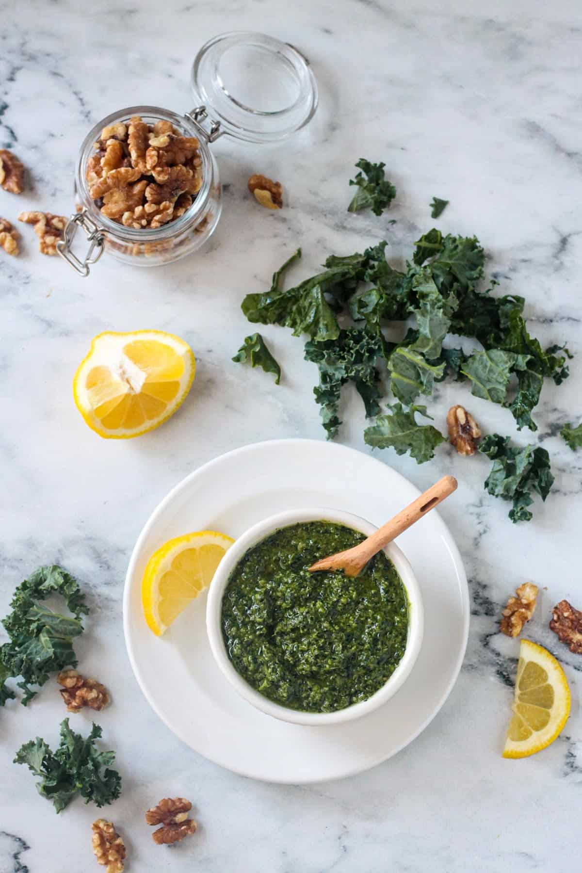 Kale walnut pesto in a white bowl next to a lemon wedge, chopped kale, and a small jar of walnuts.