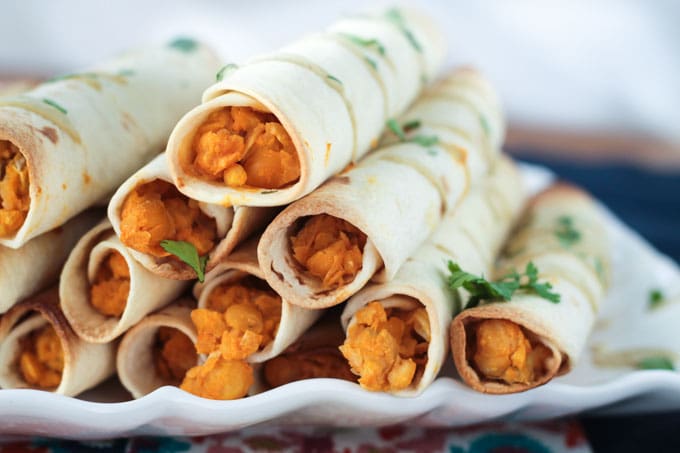 A stack of vegan taquitos, with Buffalo Chickpea Artichoke filling spilling out, on a white plate