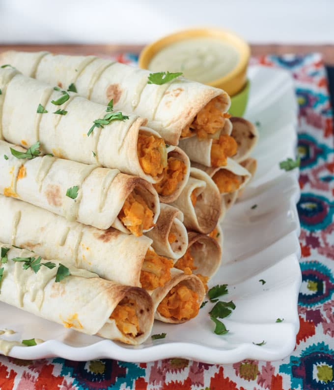 A stack of rolled tacos on a white plate, drizzled with sauce and sprinkled with fresh parsley. Extra sauce in a dipping bowl in the background.