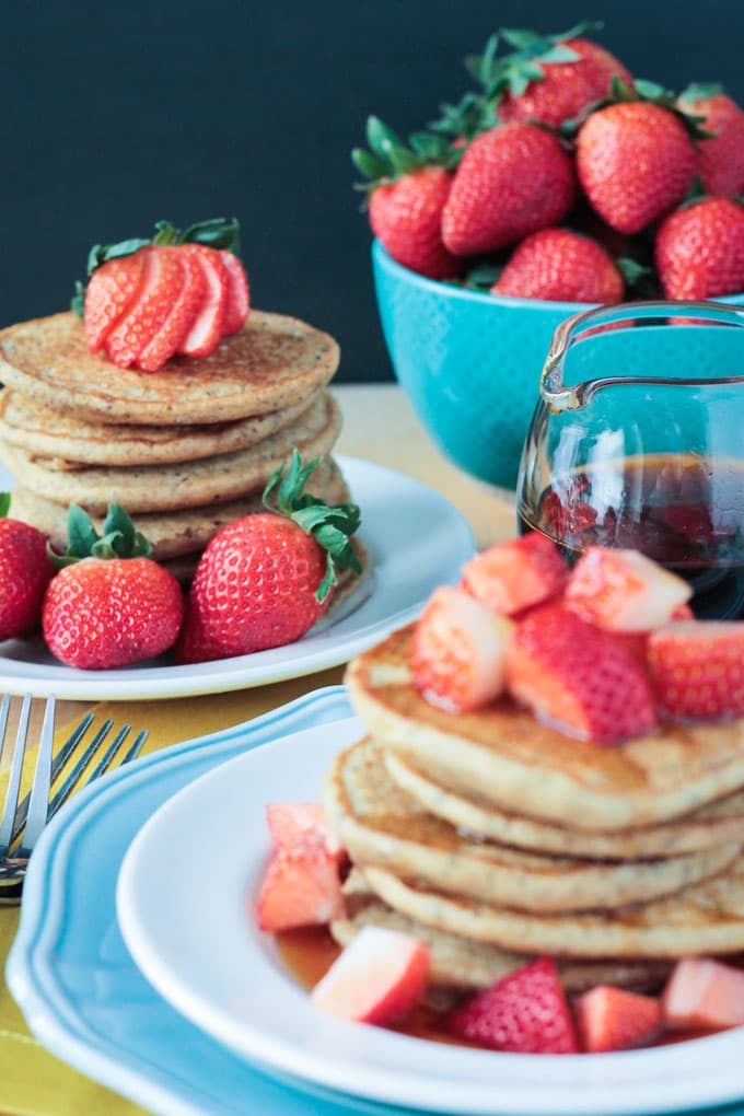 Whole fresh strawberries on a plate with a stack of lemon poppyseed pancakes. A blue bowl of fresh strawberries next to the plate.