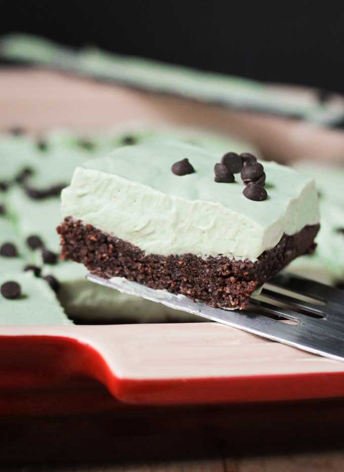 Mint Cream Icebox Dessert w/ Raw Brownie Crust - this easy, no bake dessert is loaded with nutrients thanks to vegan superfood protein powder. An indulgent dessert that is actually healthy? Yes please!! The mint cream is super light and the brownie crust is incredibly fudgy. Who needs Thin Mints when you can have Mint Cream Icebox Dessert instead? Square slice of Mint Cream Icebox Dessert on a metal spatula
