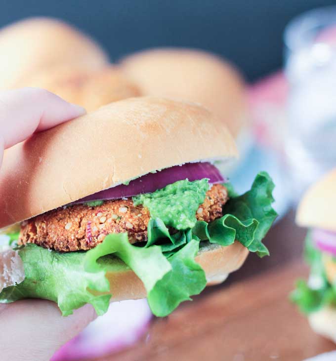 Hand holding a veggie burger topped with red onion slices, lettuce, and herbed bean spread.