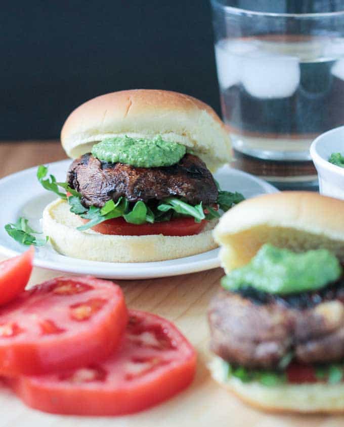 Portobello slider on a bun on a white plate topped with green pesto. Tomato slices in front of the plate.