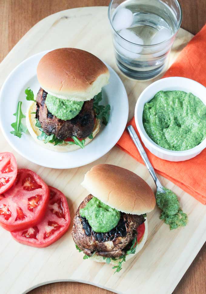 One portobello slider on a plate, one portobello slider on the table in front of the plate. Fresh tomato slices next to the burger on the table. Bowl of pesto next to the burger on the plate. Glass of ice water behind.