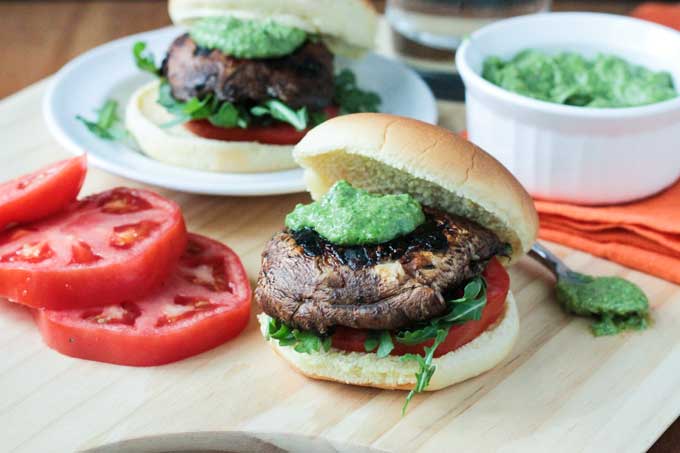 Portobello slider topped with pesto and sitting on a bed of arugula and fresh tomato slices all on a burger bun. Tomato slices next to the burger.