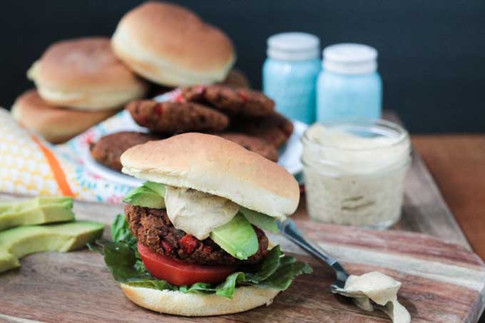 Taco Burger piled high on a bun with lettuce, tomato, avocado, and southwest ranch dressing. Small glass jar of ranch dressing behind next to a plate of burgers and buns.