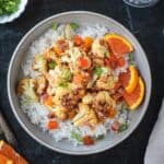 Vegan orange cauliflower in a sticky sauce with diced peppers over rice in a gray flat bowl.