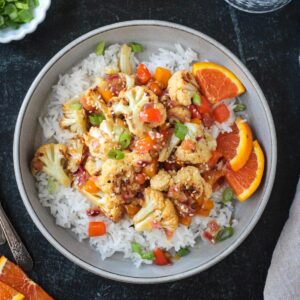 Vegan orange cauliflower in a sticky sauce with diced peppers over rice in a gray flat bowl.