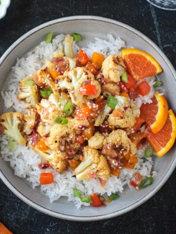Vegan orange cauliflower in a sticky sauce with diced peppers over rice in a gray flat bowl.