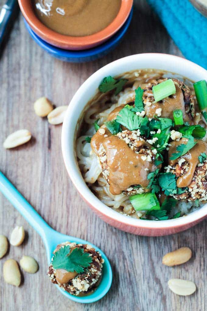 Bowl of quinoa meatballs over rice noodles next to a blue spoon holding one meatball topped with peanut sauce and a sprig of cilantro.