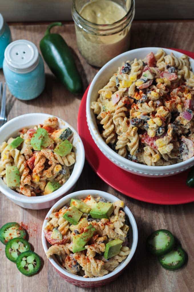 Three bowls of Southwest Black Bean Pasta Salad. Fresh jalapeño slices on the table around the bowls. Glass jar of southwest ranch dressing behind next to a full jalapeño and a salt shaker.