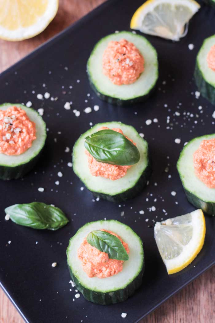 Overhead view of Cucumber Bites w/ Sun Dried Tomato Spread topped with basil and sprinkle with hemp hearts. Lemon wedges nearby.