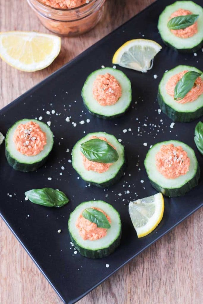 Cucumber Bites w/ Sun Dried Tomato Spread topped with small basil leaves on a black serving tray. 