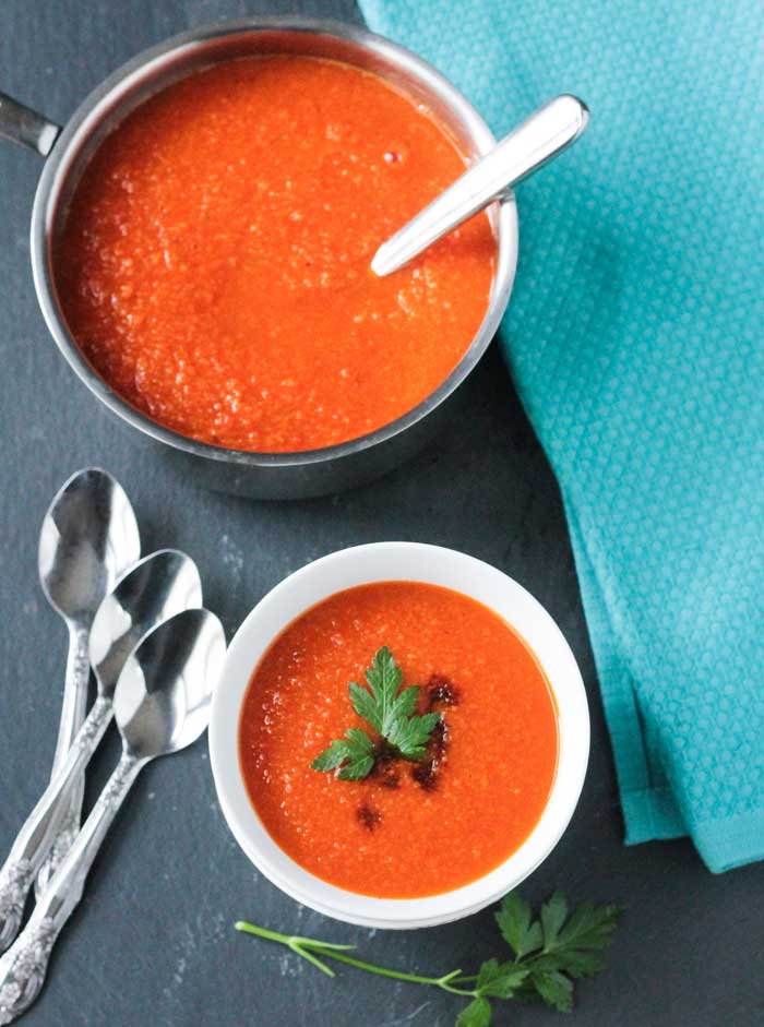 Vegan Carrot Soup in a white bowl in front of a pot of carrot soup with a metal soup ladle in it. Three metal spoons lie to the left of the bowl.
