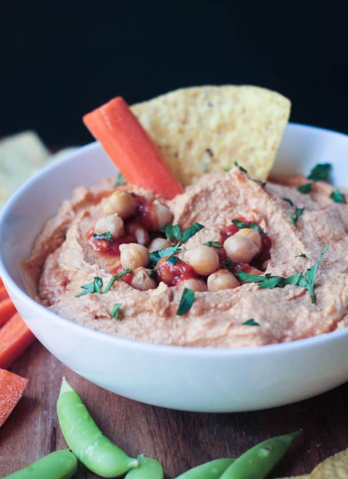 A tortilla chip & carrot stick dipped into a bowl of spicy salsa hummus.