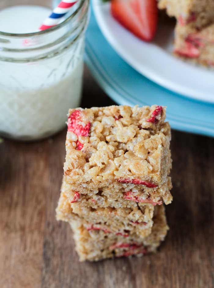 Overhead view of a stack of Brown Rice Crispy Treats w Strawberries. White plate with more rice crispy treats sits on top of a blue plate in the background next to a glass of milk. 