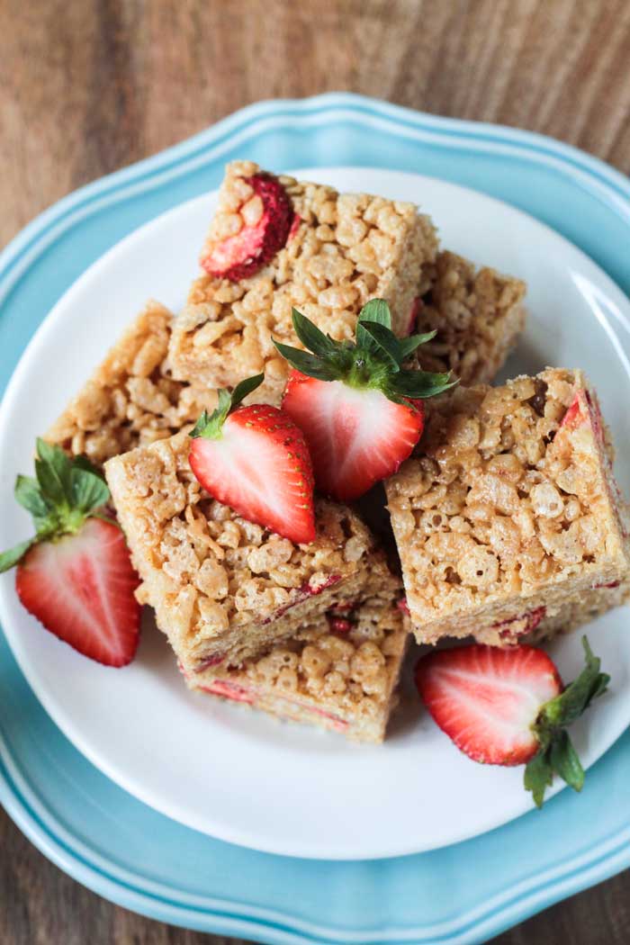 White plate with a pile Brown Rice Crispy Treats w Strawberries on top of a blue plate. Fresh halved strawberries are scattered on top.