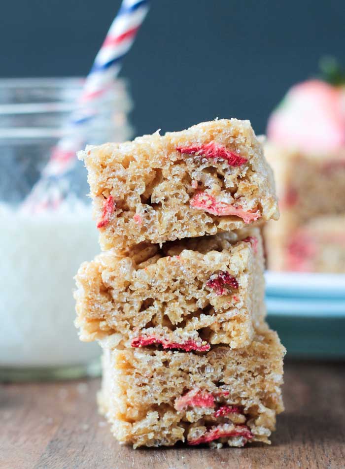 Stack of three Brown Rice Crispy Treats w Strawberries on a wooden cutting board. Glass of milk with a red, white, and blue striped straw behind.