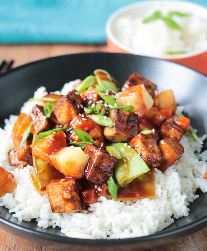 Bite size chunks of baked tofu, pineapple, snow peas, carrots, and scallions over white rice in a black bowl.