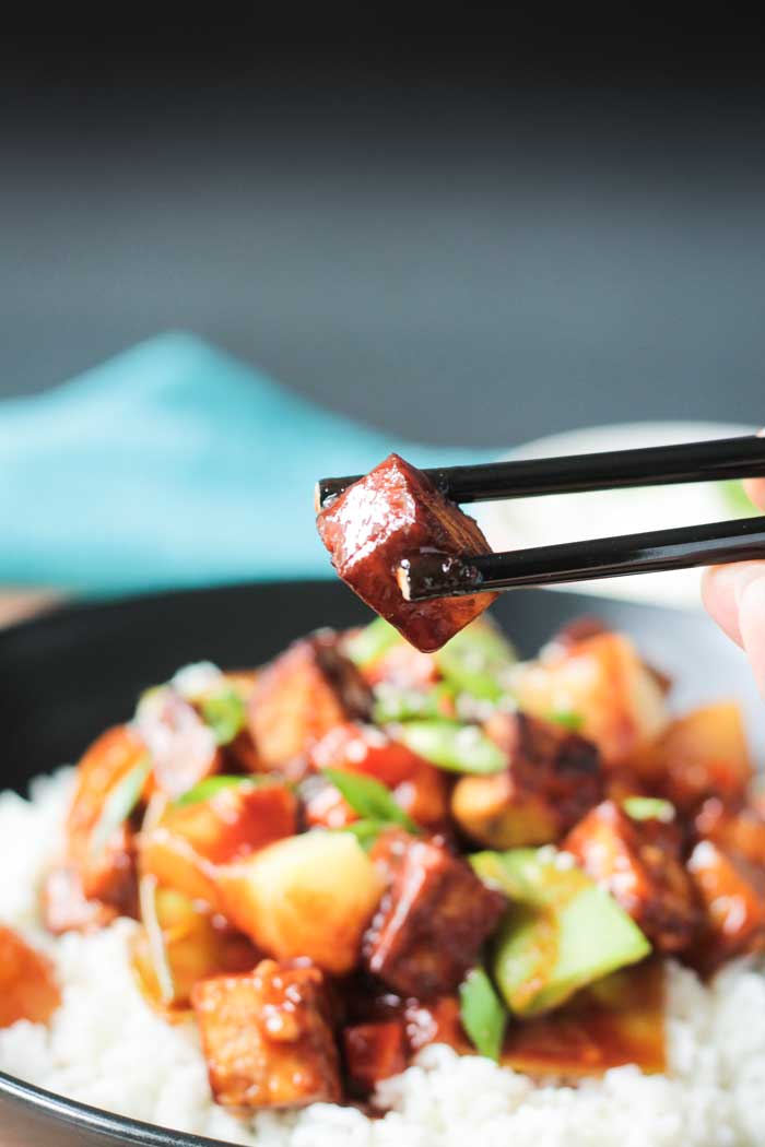 Black chopsticks holding a bite size piece of baked tofu coated in stir fry sauce over a bowl of pineapple stir fry and white rice.