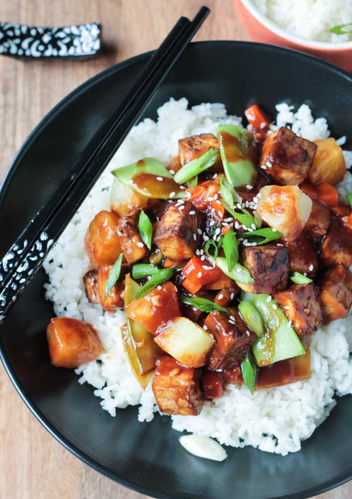 Stir Fry of baked tofu, pineapple, carrots, snow peas, and scallions over white rice in a black bowl. Black & white chopsticks lie across the top of the bowl. 