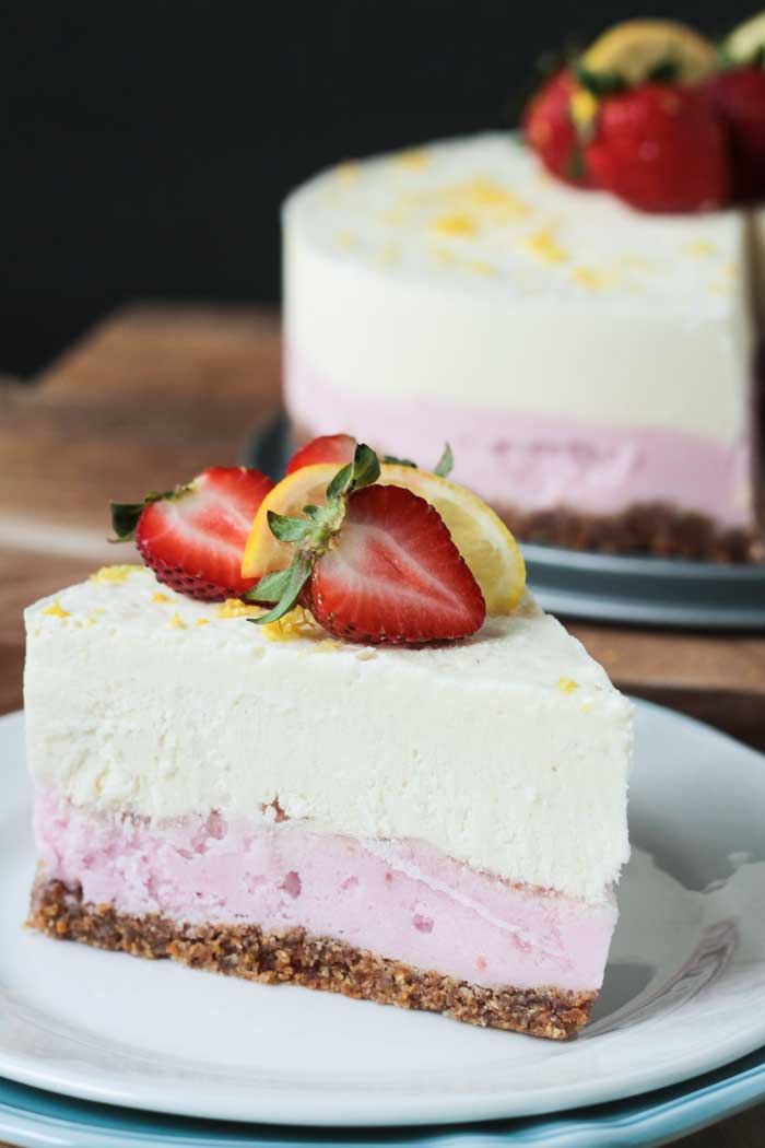 One slice of ice cream cake showing layers of pretzel crust, strawberry ice cream, and lemonade ice cream. Garnished with fresh berries and lemon. The rest of the cake in the background.