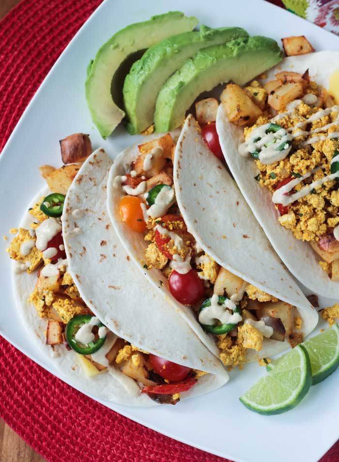 A white plate on a red placemat featuring Healthy Breakfast Tacos with scrambled tofu, roasted potatoes, jalapeños, cherry tomatoes, and avocado, with a drizzle of sauce. Lime wedges on the side.