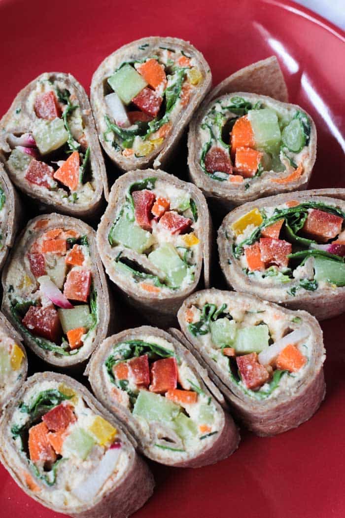 Tortilla Rollups w/ Dairy Free Veggie Cream Cheese - These creamy, crunchy little bites are the perfect appetizer or lunch. Loaded with dairy free veggie cream cheese, then topped with more fresh cut veggies, and all rolled up into a tortilla and cut into bite size pieces - these are not only delicious, but so fun to eat! Perfect for your child's lunchbox - customize it with their favorite veggies. Veggie Tortilla Rollups arranged in a circle on a red plate.