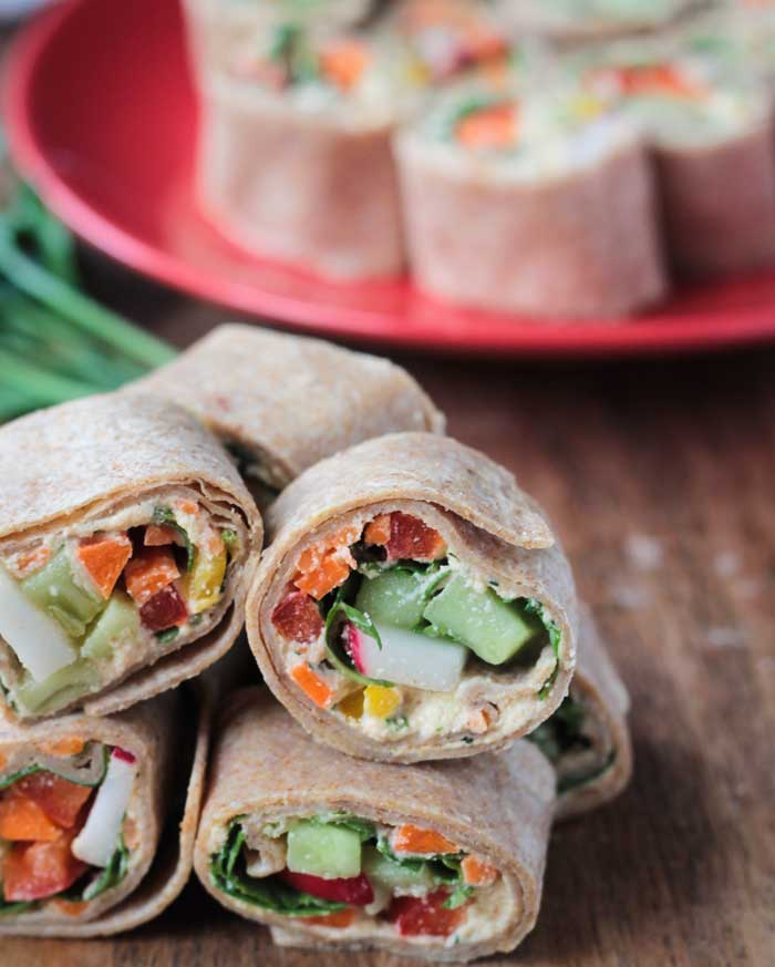 Tortilla veggie rollups stacked up on a cutting board. Red plate of more rollups in the background.