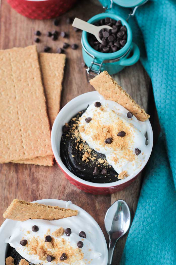 S'mores Pudding Bowl with dairy free whipped topping in small red/white bowl. Graham crackers next to the bowl. Small blue bowl of mini chocolate chips behind. Blue dish cloth to the side.