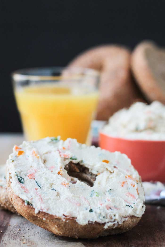 Dairy Free Veggie Cream Cheese Spread on a bagel half. Glass of orange juice and more bagels in the background.