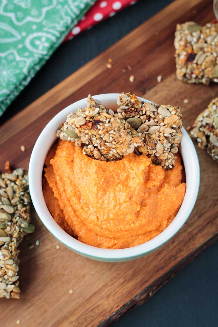 Bowl of red pepper hummus with two seed crackers dunked into one side.
