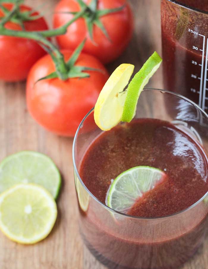 Spicy Tomato Juice in a glass with a lime slice floating on top. One lime slice and one lemon slice on the edge of the glass.