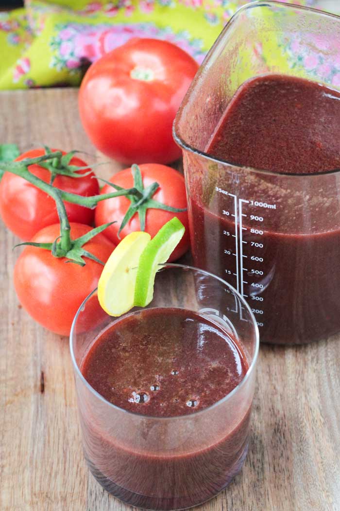 Spicy Tomato Juice in a glass garnished with a lemon and lime slice on the rim. Behind is a pitcher of more juice and a pile of fresh tomatoes.