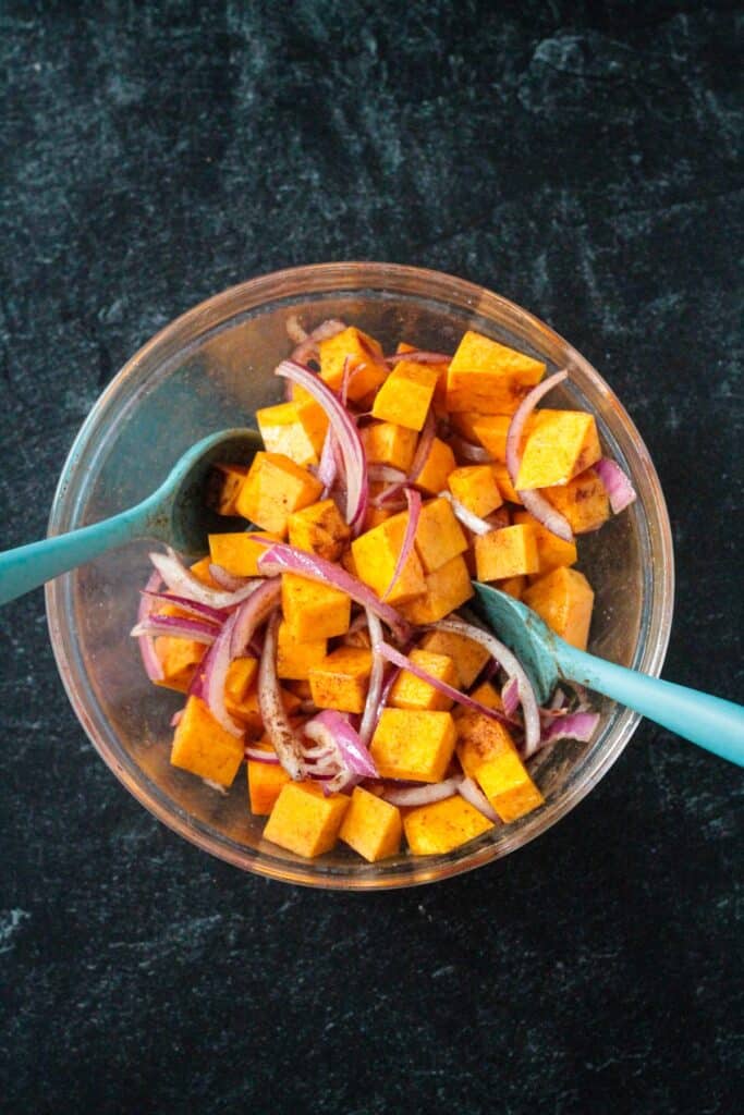 Raw cubed squash and sliced onions in a bowl mixed with oil and spices.